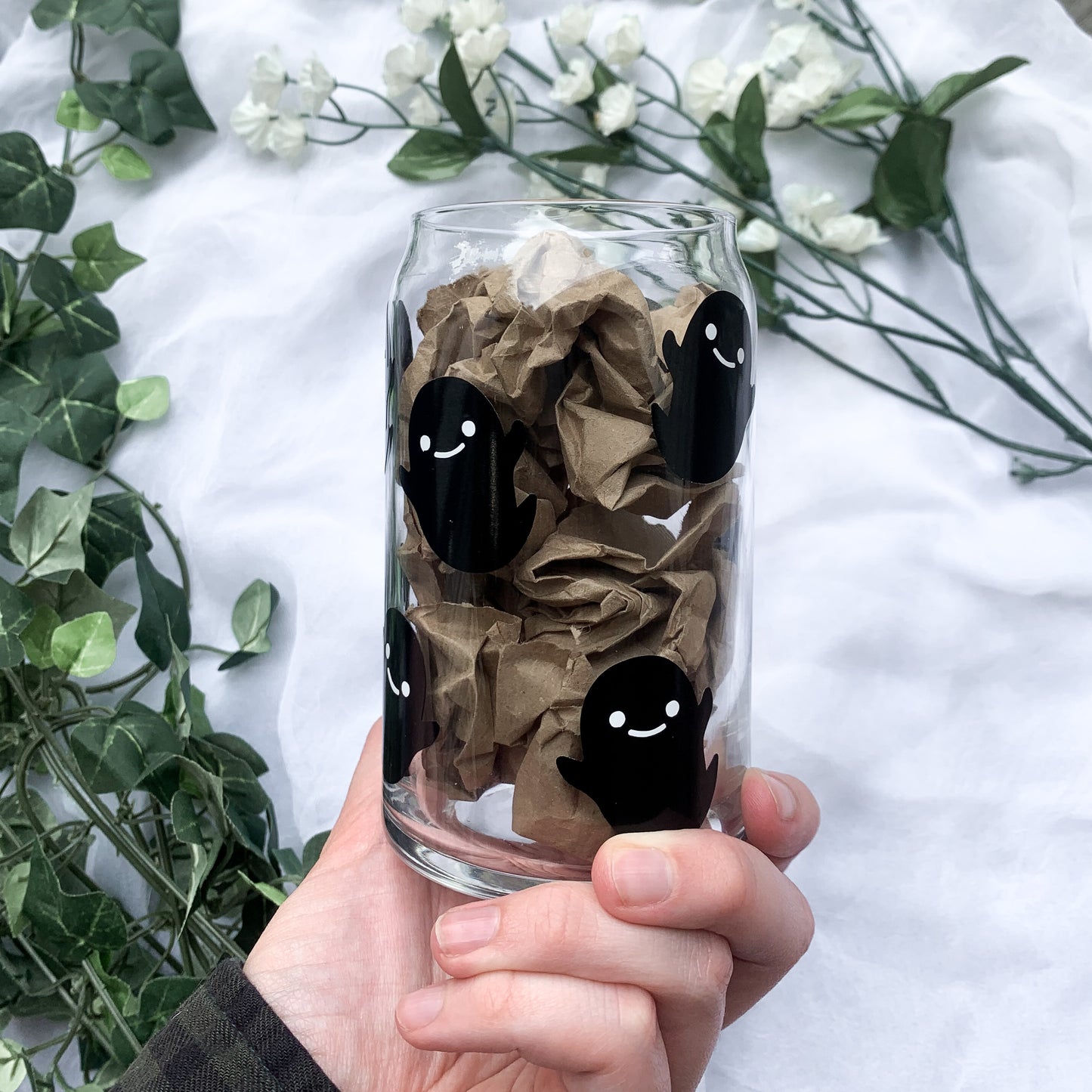 A hand holding the side of the glass cup that shows only the cute spooky ghosts with brown crinkled paper filling the glass.