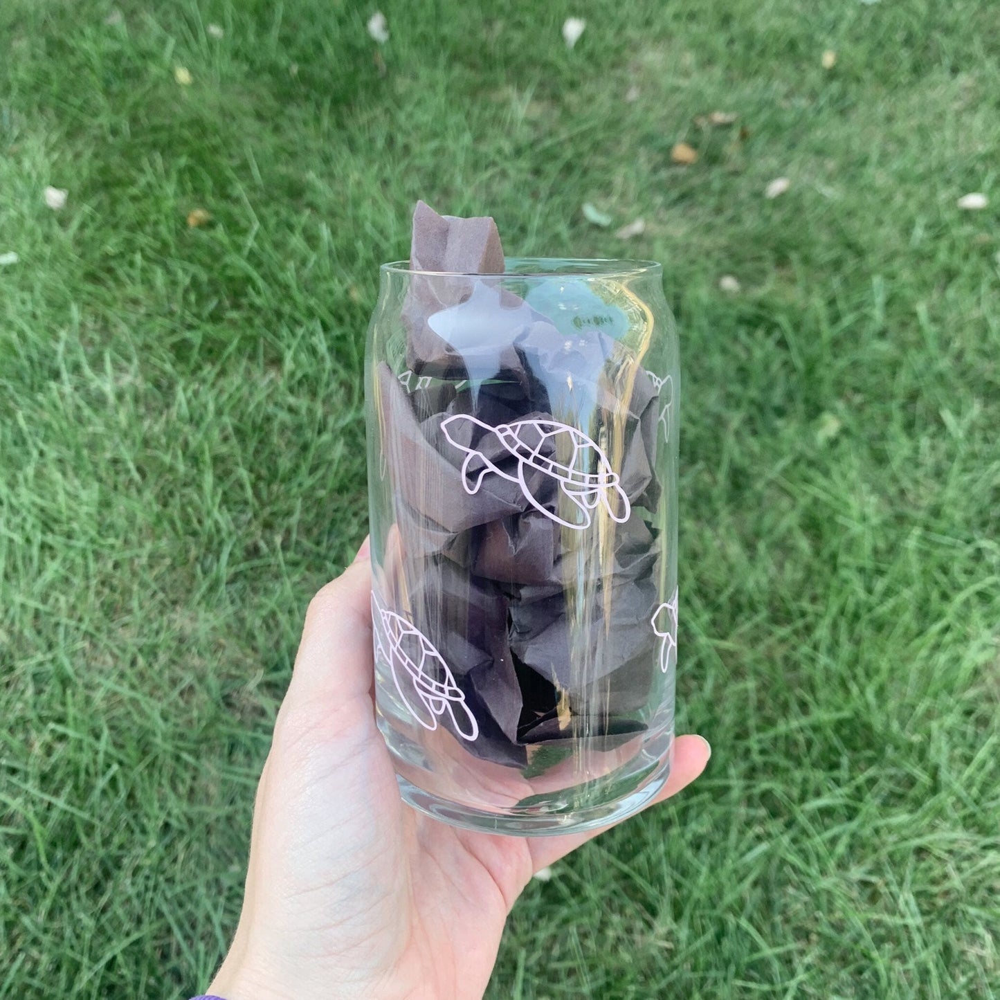 A hand holding the sea turtle glass stuffed with black crinkled paper above green grass.