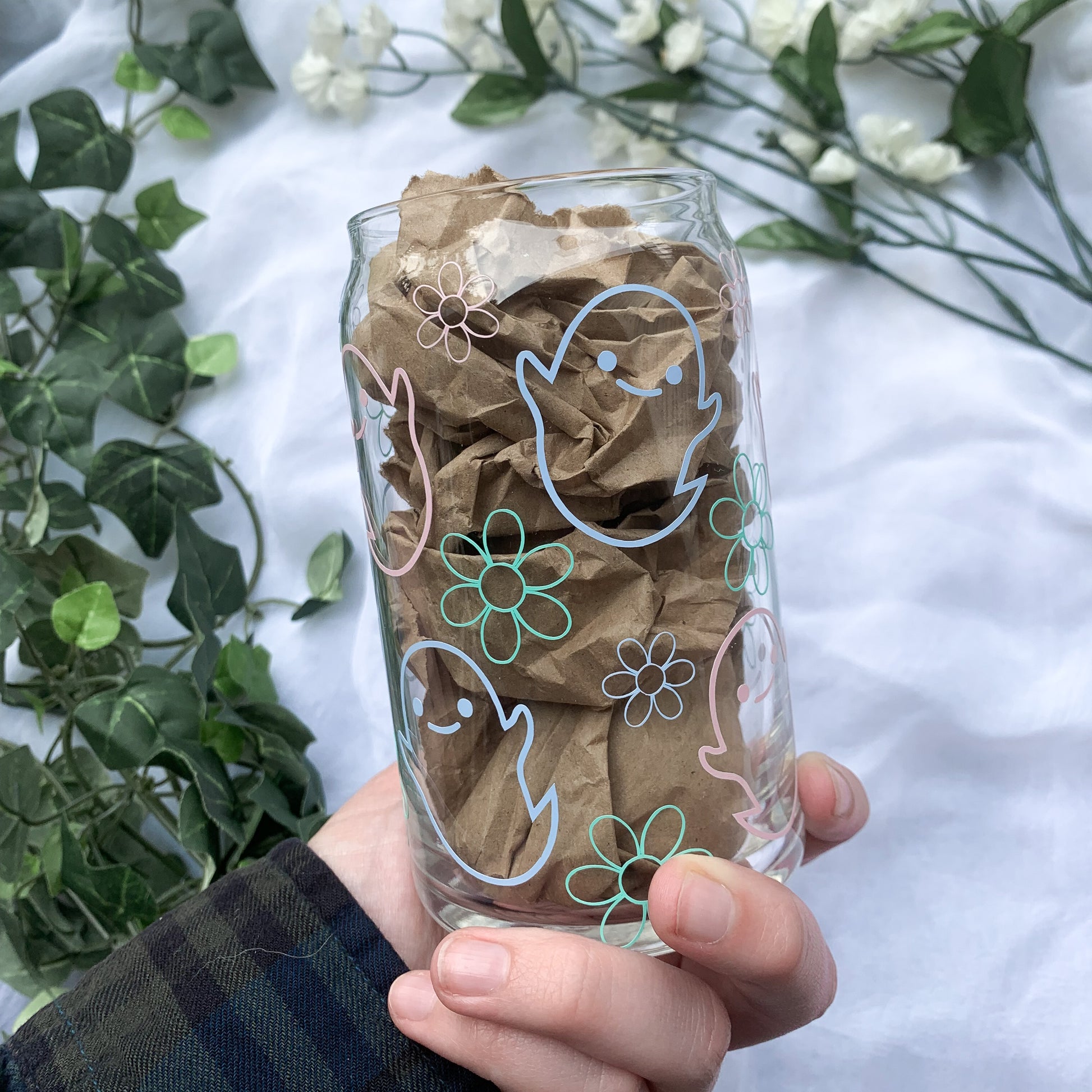 A hand is holding a glass cup designed with smiling ghosts and florals that brings positive vibes this spooky season. 